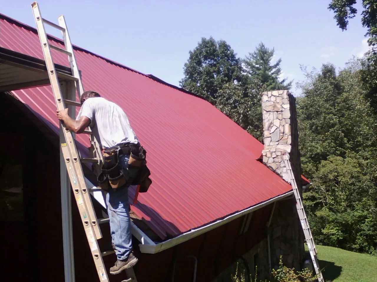 Roofer on a ladder beside a red metal roof and stone chimney — Soffit & Fascia Repair by Roofing Brothers in Malaga